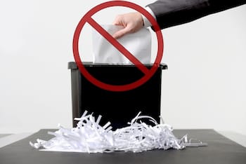 Hand of person putting a document in paper shredder with pile of previously shredded paper and a do not enter warning on it.