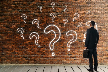 Businessman looking and thinking in front of a brick wall with question mark icons painted on it in various sizes