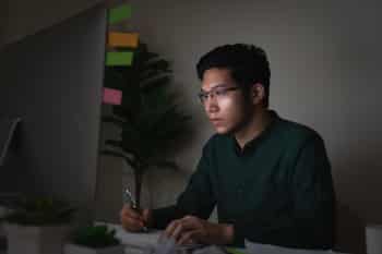 A person working on a desktop computer in a dark office. Light from the screen illuminates their face.