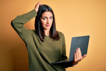 A person holding an open laptop standing with hand on head in confusion