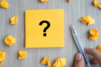 A pad of sticky notes with a large question mark on it. Multiple crumpled up sticky notes spread around.