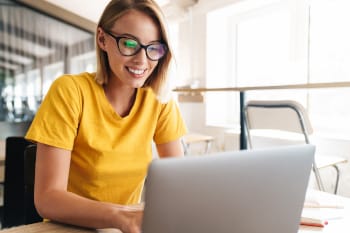 A smiling person working on a laptop in an office space.
