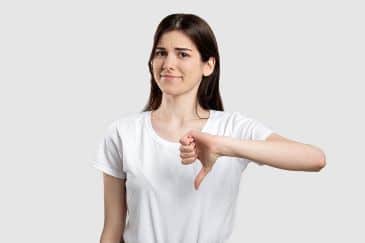 A young woman in a white t-shirt has a smirk on her face and is giving a thumbs down sign.